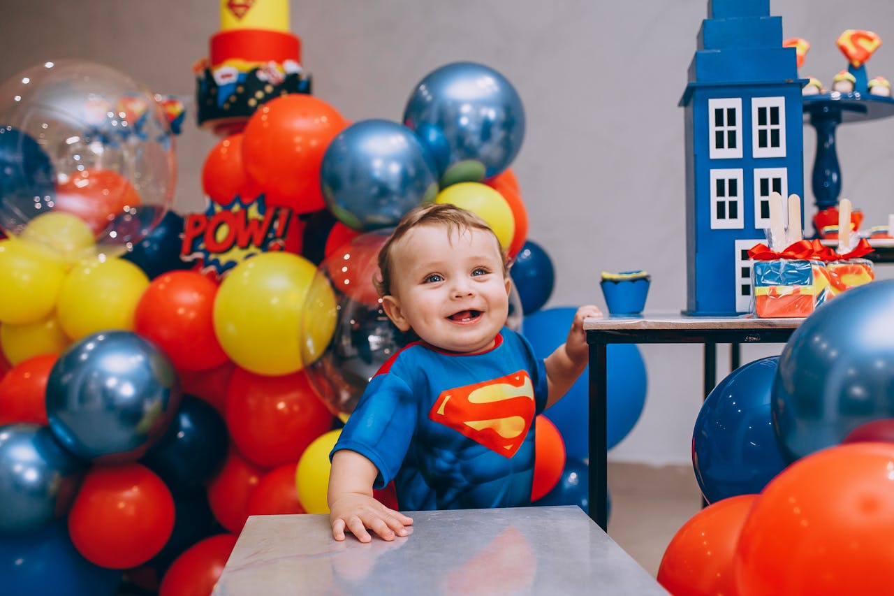 A joyful baby dressed as Superman amidst colorful superhero-themed balloons at a party.