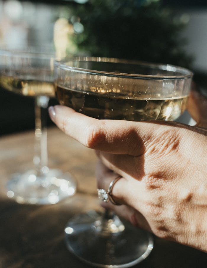 A hand holding a champagne coupe ringed with an engagement ring beside another glass on a sunlit table.